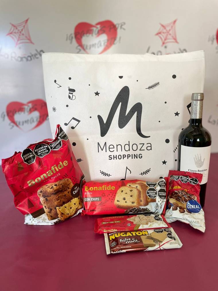 Bolsa de friselina navideña con logo de Mendoza Shopping, junto a pan dulce, budín, turrón, chocolates y una botella de vino